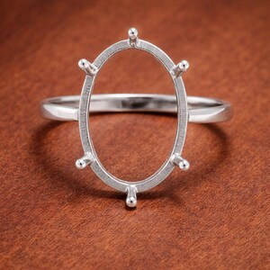 May include: A silver ring with an oval gemstone setting. The ring features six prongs around the oval, designed to hold a stone. The band is smooth and simple, and the ring is set against a brown background.