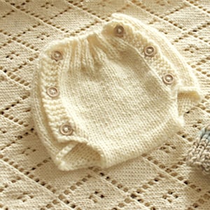 Könnte beinhalten: Ein weißer gestrickter Baby-Strampler mit Knöpfen. Der Strampler ist aus weichem, leichtem Garn gefertigt und hat ein schlichtes, klassisches Design.