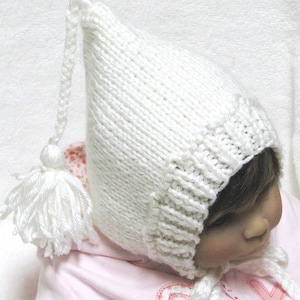 May include: White knitted baby bonnet with a long, pointed top and a white tassel. The bonnet is worn by a baby with brown hair.