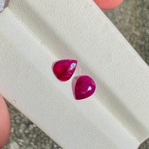 May include: Two loose, pear-shaped rubies with a deep red colour and faceted surfaces. The gemstones are positioned on a white, textured display card, showcasing their translucent quality and reflective properties under bright lighting conditions.