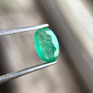 May include: A vivid green oval emerald gemstone is held by silver tweezers. The gem shows internal inclusions and facets, reflecting light. The background is blurred, highlighting the emerald's colour and clarity. A close-up view of a precious stone.