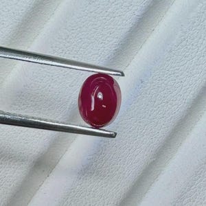 Può includere: Una gemma cabochon di colore rosso intenso e forma ovale, tenuta da una pinzetta metallica color argento. La pietra presenta una superficie liscia e lucida con un riflesso di luce visibile, che mette in risalto il colore ricco e la forma.