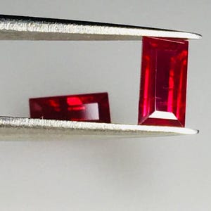 Könnte beinhalten: Zwei rechteckige, facettierte, tiefrote Edelsteine werden von einer Pinzette vor einem weißen Hintergrund gehalten. Der größere Edelstein steht aufrecht, während der kleinere horizontal liegt. Die Edelsteine haben ein brillantes, poliertes Aussehen.