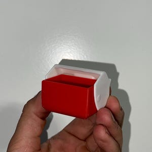 May include: A small, red and white plastic container with a hinged lid. The container is rectangular with rounded corners. The lid is white and has a curved design. The container is held in a person's hand.