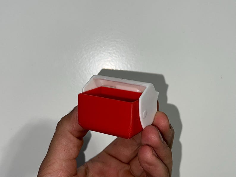 May include: A small, red and white plastic container with a hinged lid. The container is rectangular with rounded corners. The lid is white and has a curved design. The container is held in a person's hand.