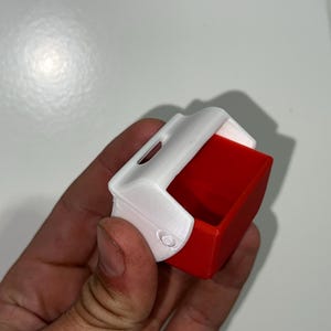 May include: A small, rectangular container with a white top and a red interior. The container has a curved top and a small opening. The object is held in a hand.