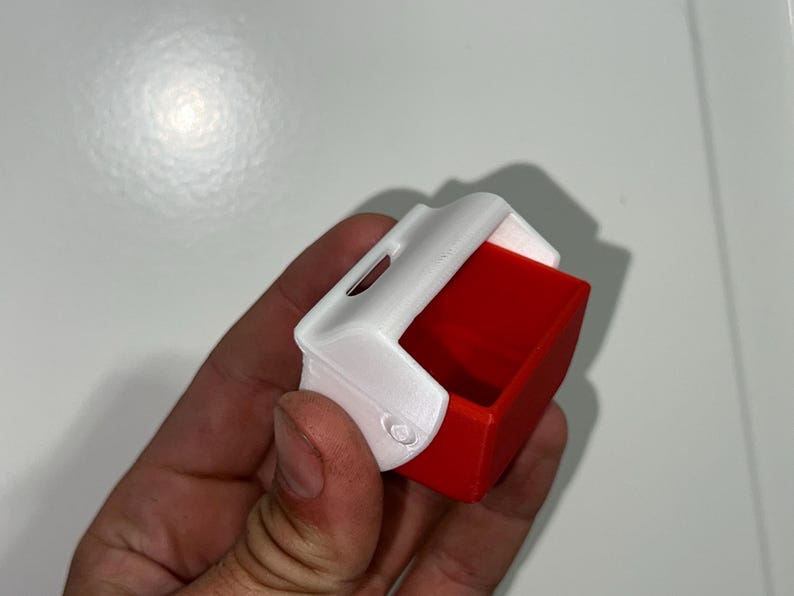 May include: A small, rectangular container with a white top and a red interior. The container has a curved top and a small opening. The object is held in a hand.