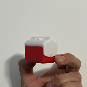 May include: A miniature red and white cooler with a handle and a latch. The cooler is held in a person's hand against a white background. The cooler is a small, detailed replica.