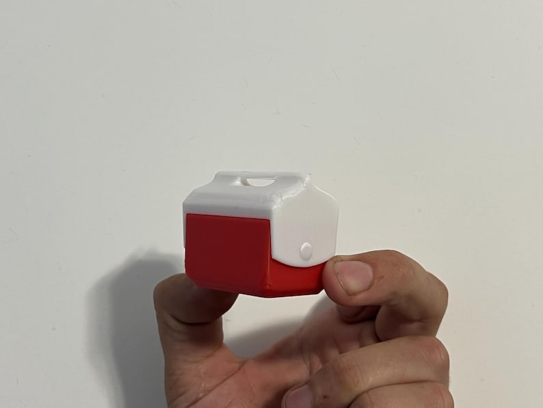 May include: A miniature red and white cooler with a handle and a latch. The cooler is held in a person's hand against a white background. The cooler is a small, detailed replica.
