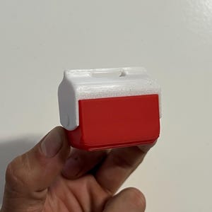 May include: A miniature red and white cooler, likely a toy or decorative item. The cooler has a rectangular red body with a white lid and handle. The design mimics a classic picnic cooler.