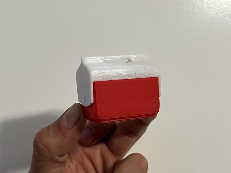 May include: A miniature red and white cooler, likely a toy or decorative item. The cooler has a rectangular red body with a white lid and handle. The design mimics a classic picnic cooler.