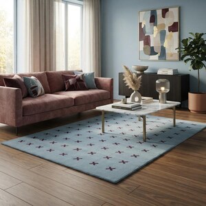 Puede incluir: Una alfombra rectangular con una base de color azul claro y un patrón repetitivo de pequeños símbolos de cruz en color granate oscuro. La alfombra está colocada sobre un suelo de madera frente a un sofá rosa y una mesa de centro de mármol blanco.
