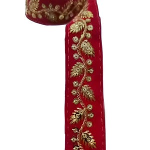 May include: Red decorative ribbon with gold floral embroidery. The ribbon is rolled up at the top and unrolled at the bottom, showcasing the detailed gold design. This trim is suitable for various craft and sewing projects.