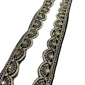 May include: Two strips of black lace with a scalloped edge and gold embroidery. The gold embroidery features a floral design with small, circular accents. The lace is against a white background.