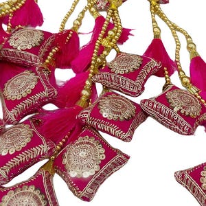 Könnte beinhalten: Nahaufnahme von dekorativen Quasten in leuchtendem Fuchsia. Jede Quaste hat ein quadratisches, besticktes Kissen mit goldenen Verzierungen und eine passende fuchsiafarbene Fransen. Die Quasten sind mit goldfarbenen Perlensträngen aufgereiht.