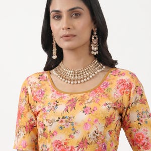 May include: A short-sleeved blouse featuring a vibrant yellow background with a multi-colored floral print in shades of pink, orange, and green. The garment has a round neckline and is paired with a gold-toned necklace and matching drop earrings.