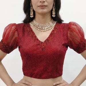 May include: A deep red blouse featuring a V-neckline with intricate beadwork, short sheer puff sleeves, and a subtle tonal floral pattern. The garment is paired with a cream-coloured skirt and a gold-toned necklace and earring set.