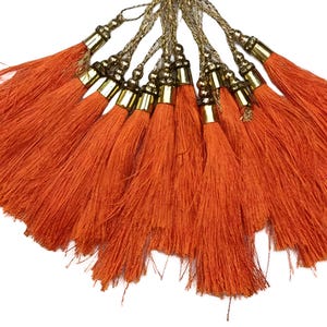 Könnte beinhalten: Eine Ansammlung orangefarbener Quasten mit goldfarbenen Metallkappen und einer goldfarbenen geflochtenen Kordel. Die Quasten bestehen aus einem weichen, faserigen Material und sind fächerförmig angeordnet. Die Kappen haben ein dekoratives Perlendesign.