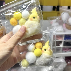 May include: Two clear plastic pouches each containing a needle-felted cockatiel figurine with a yellow crest, orange cheek patches, and black eyes, accompanied by several small, round, fuzzy pom-poms in shades of yellow and white.