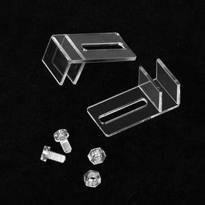 May include: Two clear acrylic L-shaped mounting brackets, each featuring a rectangular slot for adjustment. The set includes two small metal screws and two matching hexagonal nuts, providing the necessary hardware for installation and secure fastening.