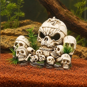 May include: An aquarium ornament featuring a large central skull with a small crab on top, surrounded by several smaller skulls and green plastic plants. The skulls are off-white with dark recessed eye sockets and teeth, arranged on a rocky base.