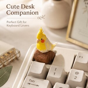 Puede incluir: Una figura de cacatúa ninfa amarilla y blanca posada sobre una tecla en forma de tronco marrón, instalada en un teclado de computadora blanco. El texto "Cute Desk Companion" y "Perfect Gift for Keyboard Lovers" es visible en la imagen.