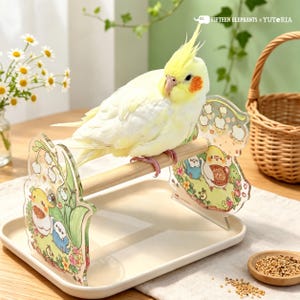 May include: A light yellow cockatiel with orange cheek patches perched on a wooden bird stand. The stand features two clear acrylic side panels decorated with colorful illustrations of small birds and flowers, set on a beige tray.
