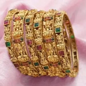 Könnte beinhalten: Ein Stapel verzierter Goldarmreifen mit aufwendigen Designs und eingebetteten roten und grünen Edelsteinen. Die Armreifen sind auf einem weichen, rosa Stoff angeordnet und zeigen ihre detaillierte Handwerkskunst.