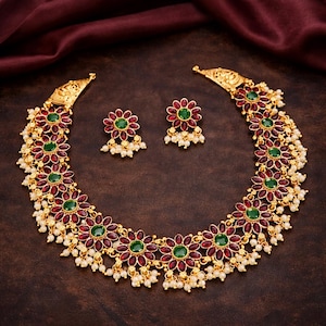 May include: A gold-toned necklace and matching earrings set. The necklace and earrings feature a floral design with red and green gemstones, accented with small white pearls. The jewelry is displayed on a dark brown surface.