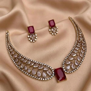 Könnte beinhalten: Ein goldfarbenes Schmuckset mit Halskette und passenden Ohrringen. Die Halskette hat einen großen, rechteckigen, rubinroten Mittelstein und ein geschwungenes Design mit klaren, tropfenförmigen Steinen. Die Ohrringe haben rubinrote Steine.