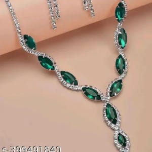 Puede incluir: Un conjunto de collar y pendientes plateados con piedras preciosas verdes esmeralda con forma de marquesa. El collar tiene un diseño de colgante en forma de Y, con las piedras preciosas engastadas en un pavé de cristales transparentes. Los pendientes tienen un diseño a juego.