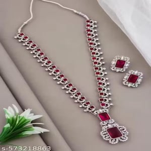 May include: A silver necklace and earring set with rectangular, deep red gemstones. The necklace has a pendant with a large red stone, and the earrings match. The set is decorated with small, clear crystals.