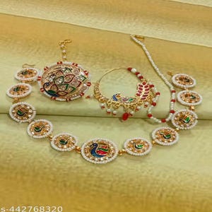 May include: A gold-coloured jewellery set with a peacock design. The set includes a headpiece, nose ring, necklace, and earrings. The peacock design incorporates red, green, and blue accents, with white beaded detailing. The jewellery is displayed on a gold-coloured surface.