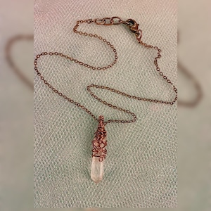 May include: A copper-colored chain necklace with a clear crystal pendant. The crystal is wrapped in copper wire. The necklace has a lobster clasp closure. The background is a light green fabric.
