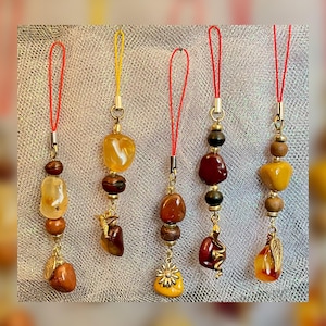 May include: Five gemstone charms with gold-colored metal accents and red or yellow cords. The charms feature various shapes and colors of stones, including amber, brown, and yellow, with decorative elements like a flower and a leaf.