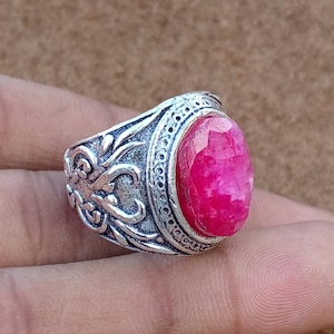 May include: A silver ring with an oval, faceted, bright pink gemstone. The ring has an ornate, engraved design around the band and the setting of the stone. The ring is shown on a brown surface.