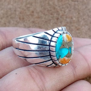 May include: A silver ring with an oval turquoise and orange stone. The ring has a detailed design on the band and around the stone. The stone is the focal point, with its vibrant colors and unique pattern. The ring is a piece of jewelry.