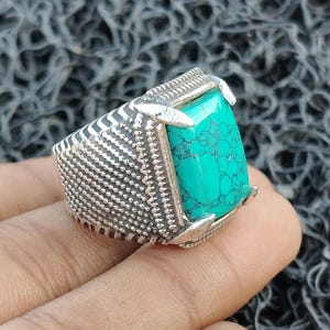 Puede incluir: Un anillo de plata con una gran piedra turquesa rectangular. El anillo tiene una banda detallada y texturizada y garras que sujetan la piedra. La piedra turquesa tiene un patrón veteado único.