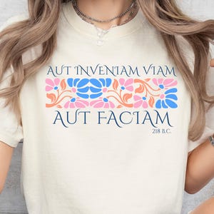 May include: Cream-colored t-shirt with the Latin phrase "Aut inveniam viam aut faciam" in navy blue. The shirt features a floral design in pink, blue, and orange. The text "218 B.C." is below the phrase.