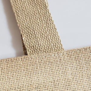 May include: Close-up of a natural-coloured burlap tote bag. The bag features a woven texture with a sturdy strap. The strap is sewn onto the bag, and the bag is made of a durable material.