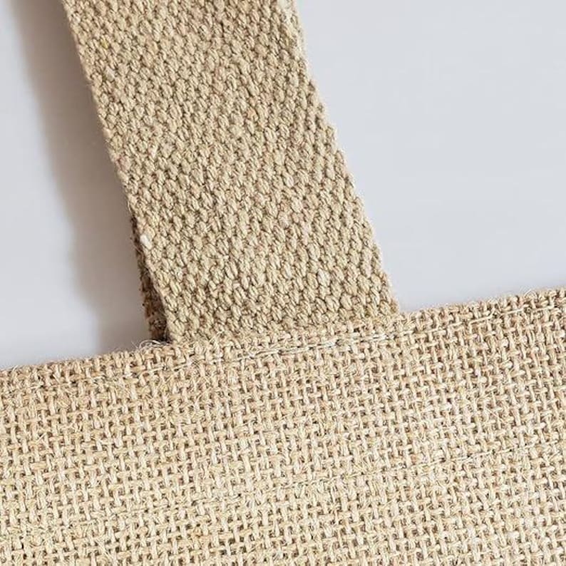 May include: Close-up of a natural-coloured burlap tote bag. The bag features a woven texture with a sturdy strap. The strap is sewn onto the bag, and the bag is made of a durable material.