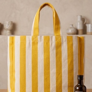 May include: A striped tote bag with yellow and white vertical stripes. The bag has yellow handles. A small brown bottle of olive oil and a bundle of lavender are in front of the bag. The items are on a wooden surface.