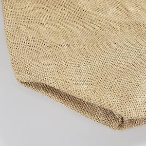 May include: Close-up of a tan burlap fabric, showing the woven texture and natural colour variations. The fabric is folded over, revealing the edge and the interior weave. The neutral tones suggest a rustic or eco-friendly theme.