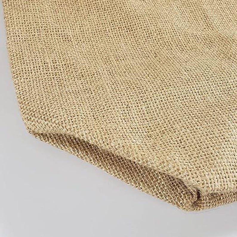 May include: Close-up of a tan burlap fabric, showing the woven texture and natural colour variations. The fabric is folded over, revealing the edge and the interior weave. The neutral tones suggest a rustic or eco-friendly theme.