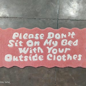 Pode incluir: Tapete retangular rosa com borda ondulada. O tapete apresenta o texto branco "Please DON'T Sit On My Bed With Your Outside Clothes". Este tapete decorativo é projetado para uso interno e mede aproximadamente 70 cm de comprimento.