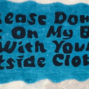 May include: A rectangular, turquoise rug with a wavy border. The rug features the phrase "Please Don't Sit On My Bed With Your Outside Clothes" in bold, black letters. The rug is made of a soft, plush material.