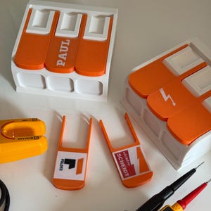 Könnte beinhalten: Eine Auswahl an elektrischen Testwerkzeugen und Zubehör. Enthält weiße und orangefarbene Halter, Messleitungen und ein gelbes Multimeter. Die Halter tragen die Aufschriften "PAUL" und "elektro SCHEIDT".