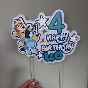 May include: A layered cake topper featuring the cartoon characters Bluey and Bingo, the number 4, and the text "HAPPY BIRTHDAY LEO". The design includes silver glitter accents, blue and orange color schemes, and two clear plastic sticks for support.