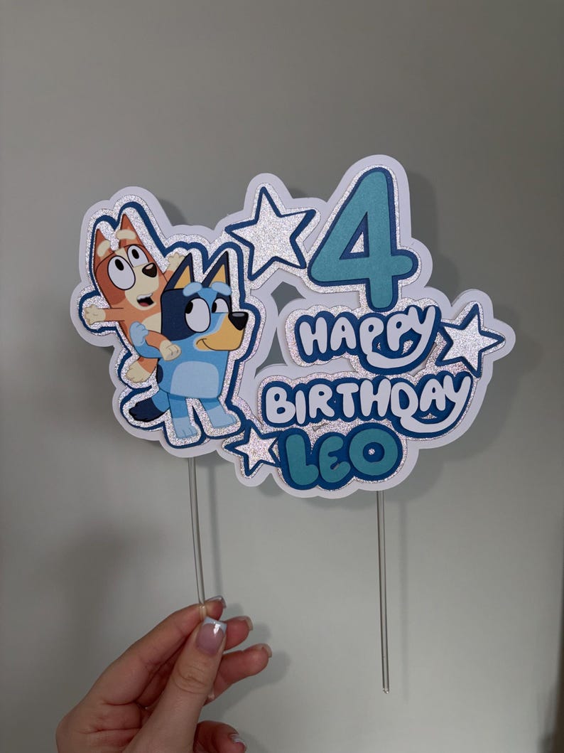 May include: A layered cake topper featuring the cartoon characters Bluey and Bingo, the number 4, and the text "HAPPY BIRTHDAY LEO". The design includes silver glitter accents, blue and orange color schemes, and two clear plastic sticks for support.