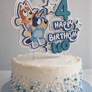 May include: A white frosted cake decorated with blue and white sprinkles, topped with a layered glittery cake topper featuring two cartoon dogs, the number 4, and the text Happy Birthday Leo in blue and silver tones with star accents.